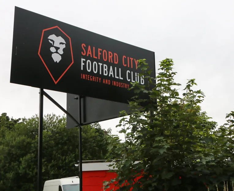 Biển hiệu lớn với logo Salford City FC tại sân Moor Lane, chào đón người hâm mộ
