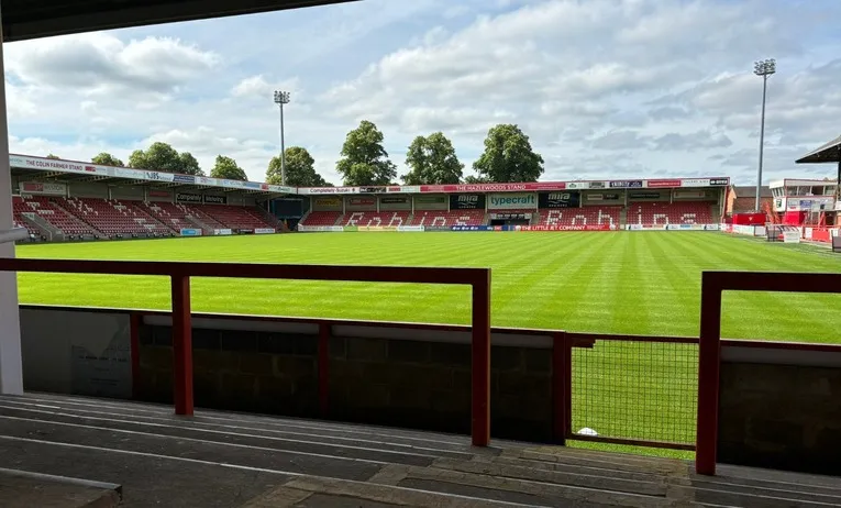 Góc nhìn từ khán đài đứng (terraces) tại sân Whaddon Road, nơi người hâm mộ cuồng nhiệt đứng cổ vũ, cảm nhận không khí trận đấu sôi động.
