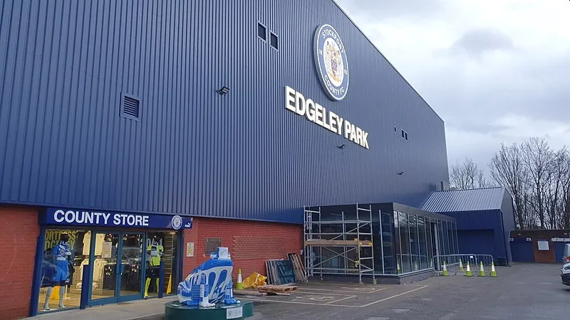 Hình ảnh mặt ngoài khán đài Cheadle End và cửa hàng bán đồ lưu niệm của Stockport County, thể hiện sự kết nối với cộng đồng.